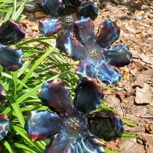 May include: A metal garden sculpture of four iridescent blue flowers with a textured finish. The flowers are arranged in a cluster and are set against a background of green grass and brown mulch.