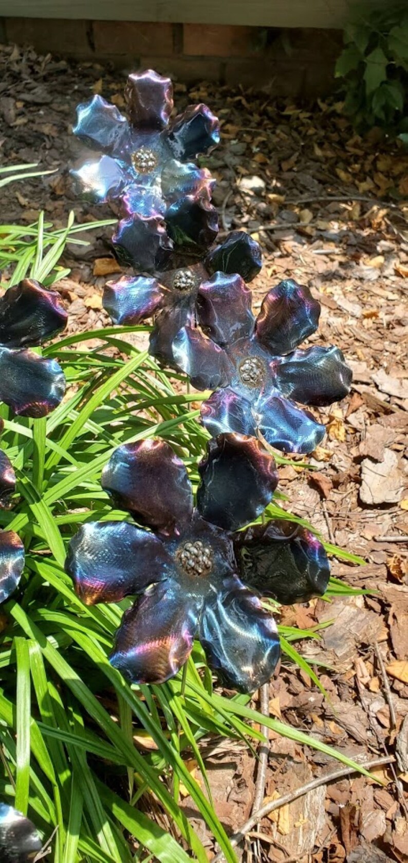 May include: A metal garden sculpture of four iridescent blue flowers with a textured finish. The flowers are arranged in a cluster and are set against a background of green grass and brown mulch.