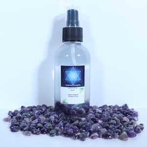 May include: A clear glass bottle with a black spray top filled with a clear liquid. The bottle has a label that says "Protection Spray" and "Against Negative Energies 2 oz/ 59 ml". The bottle is sitting on a bed of purple amethyst stones.