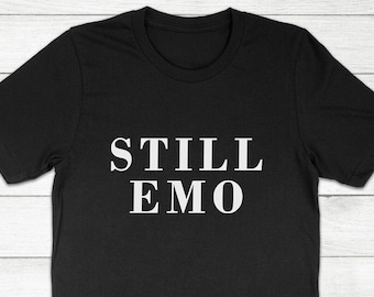 May include: Black crew neck t-shirt with the white text "STILL EMO" printed across the chest. The shirt is made of a soft material and is displayed on a light wood grain surface.