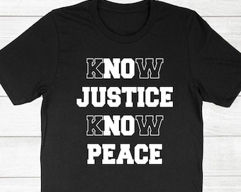 Może przedstawiać: Czarna koszulka z białym napisem "KNOW JUSTICE KNOW PEACE" ułożonym pionowo. Tekst ma pogrubioną, obrysowaną czcionkę. Koszulka jest prezentowana na jasnej powierzchni z usłojeniem drewna.