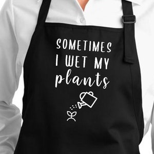 Op de afbeelding: Zwarte schort met twee voorzakken en een verstelbare nekband. De schort heeft de witte tekst "SOMETIMES I WET MY plants" met een gieter en een plantafbeelding. Gedragen over een wit overhemd.