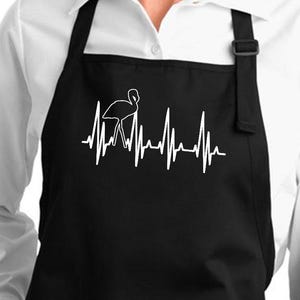 Peut inclure: Tablier noir avec deux poches avant et un motif de battement de cœur de flamant rose blanc. Le flamant rose est représenté dans la ligne du battement de cœur. Le tablier a une sangle de cou et des liens de taille noirs.