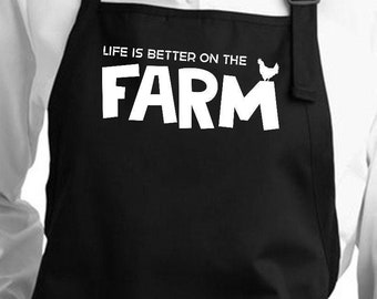 May include: Black apron with two front pockets and adjustable neck strap. The apron features the white text "LIFE IS BETTER ON THE FARM" with a small chicken graphic. The apron is worn over a white shirt.