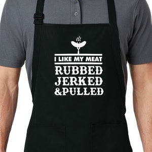 Puede incluir: Delantal negro con dos bolsillos delanteros y correa ajustable para el cuello. El delantal presenta texto blanco que dice "I LIKE MY MEAT RUBBED JERKED & PULLED" con un gráfico de salchicha. El delantal lo lleva una persona.