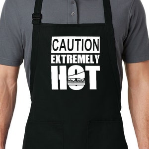 Pode incluir: Avental preto com dois bolsos frontais e alça de pescoço ajustável. O avental apresenta o texto branco "CAUTION EXTREMELY HOT" com um gráfico de grelhador. O avental é usado por uma pessoa vestindo uma camisa polo cinza.