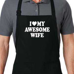 Puede incluir: Delantal negro con dos bolsillos delanteros y correa ajustable para el cuello. El delantal presenta el texto blanco "I ❤️ MY AWESOME WIFE". El delantal lo lleva una persona con una camisa polo gris.