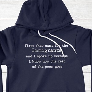 May include: A navy blue hooded sweatshirt featuring white text that reads: First they came for the Immigrants and i spoke up because i know how the rest of the poem goes. The hoodie includes a front kangaroo pocket and white drawstring cords.