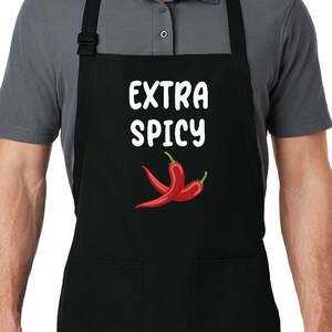 May include: Black apron with two front pockets. The apron features the text "EXTRA SPICY" in white, above a graphic of two red chili peppers. The apron is worn by a person.