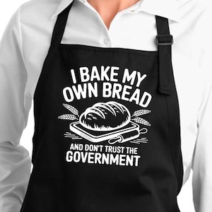 May include: A black apron featuring a white graphic of a loaf of bread on a cutting board with a rolling pin and wheat stalks. The text reads: I BAKE MY OWN BREAD AND DON'T TRUST THE GOVERNMENT. The apron has two front pockets and neck straps.