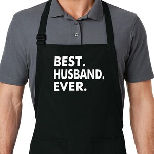 Puede incluir: Delantal negro con dos bolsillos delanteros y el texto blanco "BEST. HUSBAND. EVER." El delantal tiene una correa para el cuello y lazos en la cintura. El delantal lo lleva una persona.