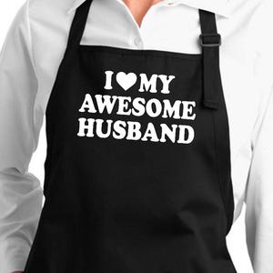 Puede incluir: Delantal negro con dos bolsillos delanteros y el texto "I ❤️ MY AWESOME HUSBAND" en blanco. El delantal se lleva sobre una camisa blanca abotonada. El delantal tiene una correa para el cuello negra y lazos negros en la cintura.