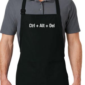 May include: Black apron with two front pockets and adjustable neck strap. The text "Ctrl + Alt + Del" is printed in white across the chest. The apron is worn by a person wearing a gray polo shirt.