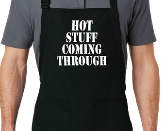 Puede incluir: Delantal negro con dos bolsillos delanteros y correa ajustable para el cuello. El delantal tiene el texto blanco "HOT STUFF COMING THROUGH". El delantal lo lleva una persona con un polo gris.