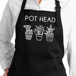 May include: Black apron featuring the text "POT HEAD" above three plant pot illustrations. The apron has two front pockets and adjustable straps. It is worn over a white shirt with a collar.