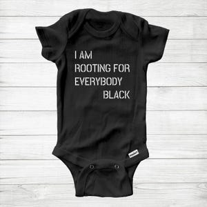 Può includere: Body da bambino nero con la scritta "I AM ROOTING FOR EVERYBODY BLACK" in lettere bianche. Il body ha maniche corte e bottoni automatici sul fondo. Lo sfondo è un legno chiaro.