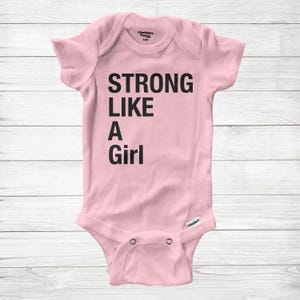 May include: A pale pink baby bodysuit featuring the black text "STRONG LIKE A Girl". The short-sleeved bodysuit has snap fasteners at the crotch. The brand name "Gerber" is printed at the top.