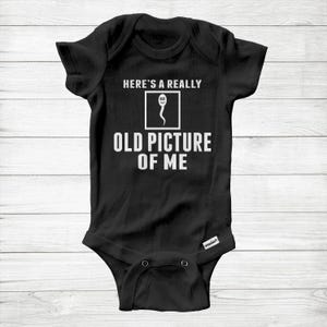 May include: Black baby bodysuit with white text: "HERE'S A REALLY OLD PICTURE OF ME." The text is above a sperm cell graphic. The bodysuit has short sleeves and press studs at the bottom. A humorous baby garment.