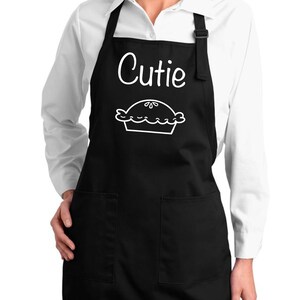May include: Black apron with two front pockets and adjustable neck strap. The apron features the word "Cutie" in white script above a white pie graphic. The apron is worn over a white collared shirt.