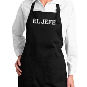 May include: Black apron with two front pockets and the text "EL JEFE" in white block letters. The apron has a black neck strap and is worn over a white long-sleeved shirt. The apron is made of a durable fabric.
