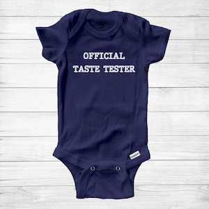 May include: Navy blue baby onesie with the text "OFFICIAL TASTE TESTER" in white block letters. The onesie has short sleeves and snaps at the bottom for easy changes. The background is a light wood grain.