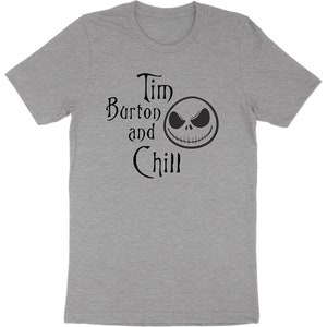 Op de afbeelding: Grijs T-shirt met de zwarte tekst "Tim Burton and Chill" en een Jack Skellington-gezicht. Het T-shirt heeft een ronde hals en is waarschijnlijk gemaakt van een zachte stof. Het ontwerp is een verwijzing naar regisseur Tim Burton.