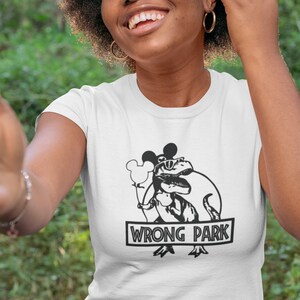 Puede incluir: Una camiseta blanca con un gr&aacute;fico en blanco y negro de un dinosaurio con orejas de Mickey Mouse y gafas de sol sosteniendo un globo. El texto "WRONG PARK" est&aacute; impreso debajo del gr&aacute;fico.