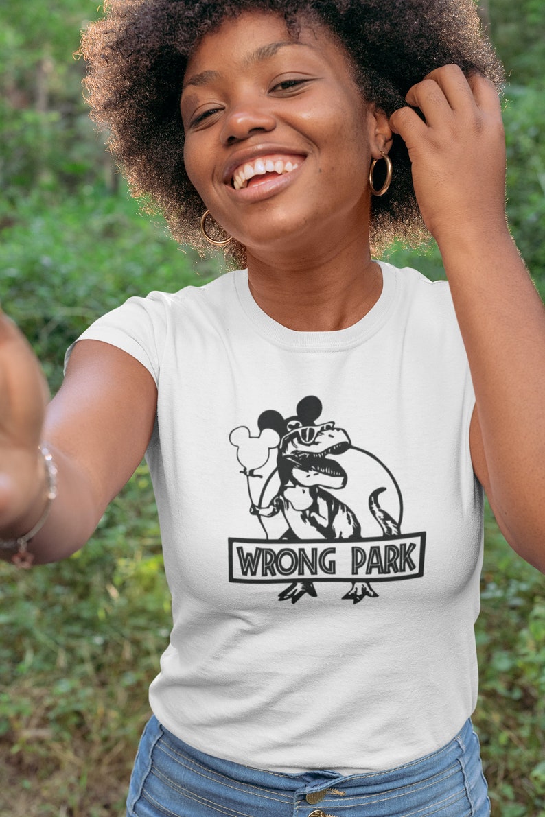 Puede incluir: Una camiseta blanca con un gr&aacute;fico en blanco y negro de un dinosaurio con orejas de Mickey Mouse y gafas de sol sosteniendo un globo. El texto "WRONG PARK" est&aacute; impreso debajo del gr&aacute;fico.