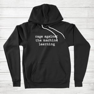 Pode incluir: Moletom preto com a frase branca "rage against the machine learning". O moletom tem capuz com cordão e um bolso frontal. O fundo é um padrão de grão de madeira clara.