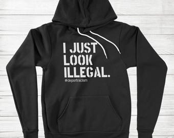 Puede incluir: Sudadera con capucha negra con texto blanco que dice "I JUST LOOK ILLEGAL." El hashtag #deportracism está debajo del texto. La sudadera tiene un bolsillo delantero y un cordón blanco.