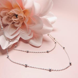 Peut inclure: Un collier en argent délicat avec de petites perles rondes est présenté sur un fond rose clair, avec une grande fleur rose clair en arrière-plan. Le collier est drapé avec grâce, mettant en valeur son élégance simple.