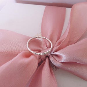 Puede incluir: Un delicado anillo de plata con una banda texturizada. El anillo se muestra en una cinta rosa.