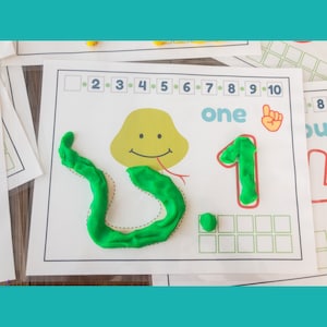 May include: A green snake made of playdough is on a white sheet of paper with a number one and the word "one" written on it. The snake is following a dotted line path. There are numbers 1 through 10 at the top of the sheet.