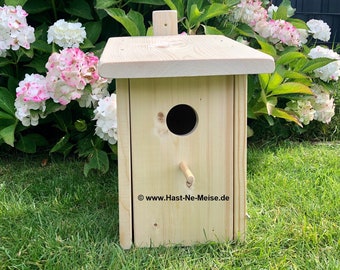 Könnte beinhalten: Ein hölzernes Vogelhaus mit einem runden Eingangsloch und einem kleinen Sitzplatz. Das Vogelhaus ist hellbraun lackiert und hat ein schlichtes Design. Das Vogelhaus steht auf einem Bett aus grünem Gras.