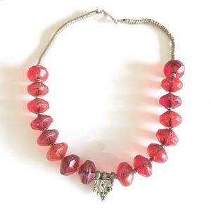 Könnte beinhalten: Halskette mit facettierten, durchscheinenden roten Perlen auf einem silberfarbenen Draht. Die Perlen variieren in Größe und Form, mit einem dekorativen Silberanhänger in der Mitte. Die Halskette wird vor weißem Hintergrund präsentiert.
