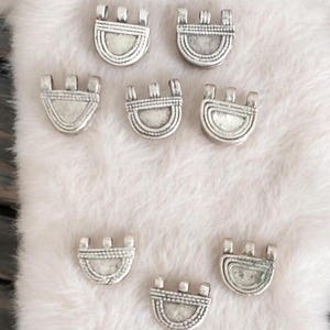 May include: Eight silver metal charms in the shape of a half-circle with a decorative design. The charms have three small loops at the top.