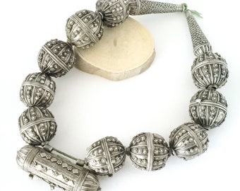 May include: A silver necklace featuring large, ornate, round beads. The necklace includes a small, rectangular pendant with a decorative design.