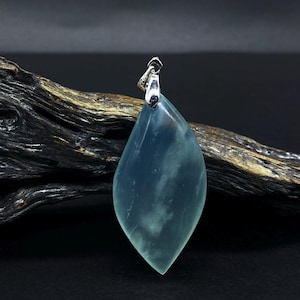 Può includere: Ciondolo a forma di goccia blu-verde traslucido con venature bianche. Ha un'ansa argentata. È esposto su un pezzo di legno scuro e invecchiato su sfondo nero. Un gioiello.