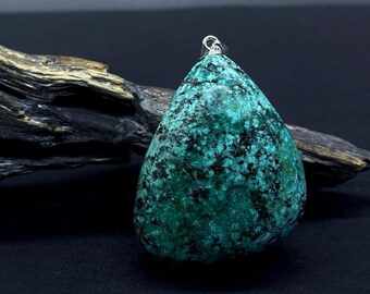 May include: A teardrop-shaped pendant with a silver bail, featuring a mottled turquoise and black stone. The pendant is set against a dark background with a piece of driftwood and the text "STEIN°ARTIG".