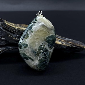 Könnte beinhalten: Ein Anhänger aus poliertem Stein mit einer silbernen Öse. Der Stein ist hauptsächlich weiß und beige mit grünen Einschlüssen. Der Anhänger wird auf einer dunklen Oberfläche mit einem Stück Treibholz im Hintergrund präsentiert. Das Wort "STEIN°ARTIG" steht unten.