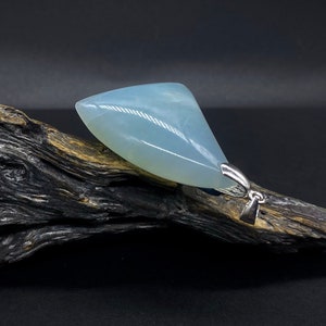 Può includere: Un ciondolo triangolare di colore blu chiaro realizzato in pietra naturale con un'anellina in argento. Il ciondolo è appoggiato su un pezzo di legno galleggiante.