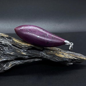 Peut inclure: Un pendentif en forme de larme de couleur violette et argentée, posé sur un morceau de bois flotté. Le pendentif a une bélière en argent et une surface texturée.