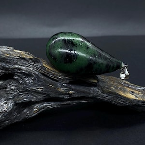 Könnte beinhalten: Ein tropfenförmiger Anhänger aus grünem und schwarzem Stein, präsentiert auf einem Stück verwittertem Holz. Der Anhänger hat eine silberne Öse und ist vor einem dunklen Hintergrund platziert. Das Wort "STEIN°ARTIG" ist unten sichtbar.