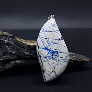 May include: A white pendant with blue and black vein-like patterns. The pendant is teardrop-shaped and hangs from a silver chain. The background is dark, with a piece of driftwood.