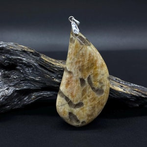 Pode incluir: Um pingente em forma de lágrima feito de pedra do sol com uma argola de prata. A pedra tem uma cor marrom clara com veios laranja e brancos.