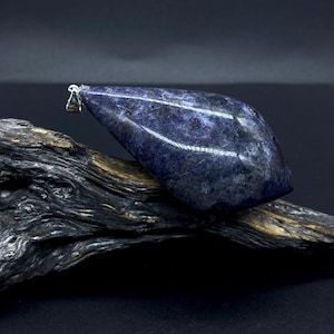 Puede incluir: Un colgante de piedra preciosa azul oscuro con una fianza plateada, apoyado en un trozo de madera flotante. La piedra tiene una superficie pulida y brillante con un patrón moteado. El fondo es de color oscuro y sólido. La palabra "STEIN°ARTIG" está en la parte inferior.
