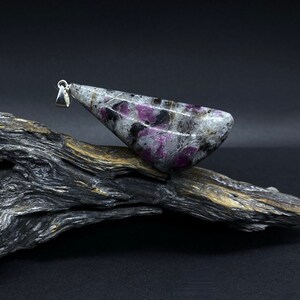 Könnte beinhalten: Ein silberner Anhänger mit einem grauen, schwarzen und rosafarbenen Edelstein mit einem einzigartigen Muster. Der Anhänger liegt auf einem Stück Treibholz.