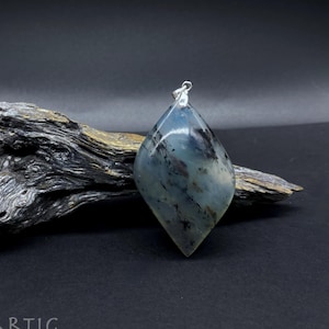 Può includere: Un ciondolo a forma di lacrima realizzato in agata grigia e nera con un'anellina in argento. Il ciondolo è appoggiato su un pezzo di legno galleggiante.
