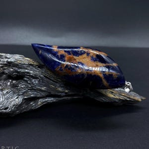 Peut inclure: Un pendentif en pierre précieuse poli et allongé, avec une bélière en argent. La pierre présente une couleur de base bleu foncé avec des veines marron et beige. Le pendentif repose sur un morceau de bois flotté gris vieilli sur fond sombre. Le texte "STEIN°ARTIG" est visible.