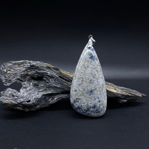 Könnte beinhalten: Ein tropfenförmiger Anhänger aus hellem grauen Stein mit dunkelgrauen und weißen Flecken. Der Anhänger hat eine silberne Öse und liegt auf einem Stück Treibholz.